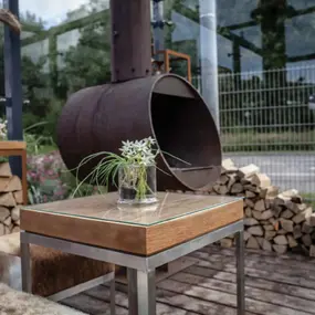 Moderner Gartentisch aus Holz und Metall mit Blumenvase vor Outdoor-Kamin und Brennholz.