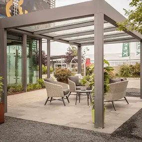 Moderne Terrassenüberdachung aus Glas mit gemütlicher Sitzgruppe und Pflanzen im Garten.